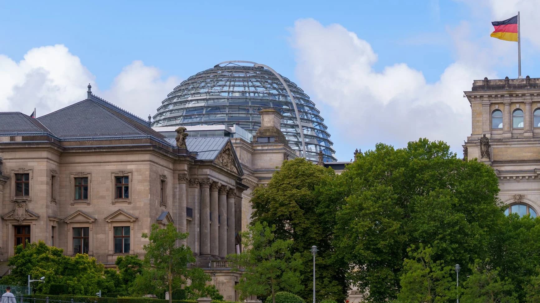 Reichstagsgebäude in Berlin mit Glaskuppel und Deutschlandfahne, davor Bäume am Spreeufer